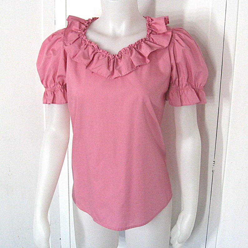 Puede incluir: Una blusa rosa de manga corta con un cuello ancho de volantes que enmarca el escote. La prenda presenta mangas abullonadas con pu&ntilde;os el&aacute;sticos de volantes y un dobladillo recto, confeccionada en un tejido liso y opaco de color s&oacute;lido.