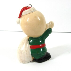 May include: A ceramic Christmas ornament of a small, bald, child-like figure wearing a green and red suit, a white Santa hat, and carrying a white sack.