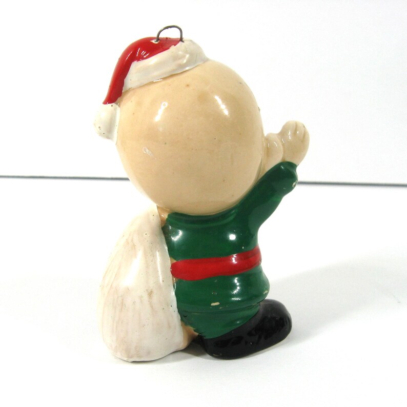 May include: A ceramic Christmas ornament of a small, bald, child-like figure wearing a green and red suit, a white Santa hat, and carrying a white sack.