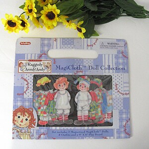 May include: A Raggedy Ann & Andy MagiCloth Doll Collection set. The package includes two magnetized dolls, eight outfits, and a 9" x 6" play board. The packaging is blue and white with a patchwork design. The dolls have red yarn hair and painted faces.