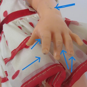 May include: A doll wearing a white dress with red polka dots and red trim. The doll's arm and hand feature several small dark spots, which are highlighted by blue arrows pointing to the specific areas of discoloration on the skin surface.