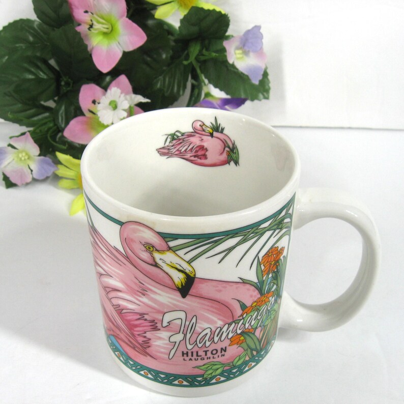 May include: White ceramic mug featuring a pink flamingo design. The mug has a green border and the text "Flamingo Hilton Laughlin" on the front.