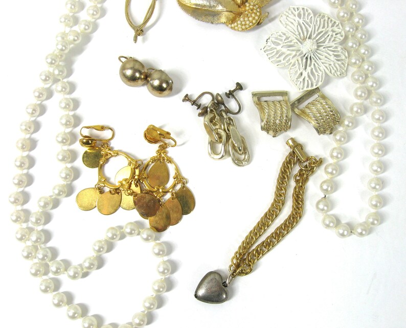 K&ouml;nnte beinhalten: Eine Sammlung von Vintage-Schmuck, darunter eine Perlenkette, goldfarbene Ohrringe, eine goldfarbene Kette mit einem silbernen Herzanh&auml;nger und eine wei&szlig;e Blumenbrosche.