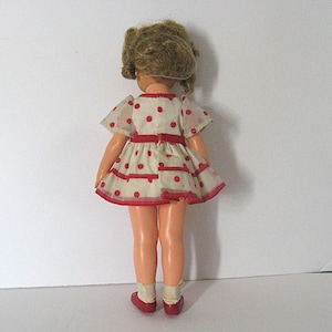 May include: A doll with blonde hair styled in curls, wearing a white short-sleeved dress featuring a red polka dot pattern and red trim at the waist and hem. The doll has light-toned skin, wears white socks, and dark red shoes, viewed from the back.