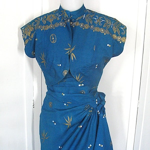 May include: A teal dress and bolero jacket set. The garments are adorned with gold leaf and geometric patterns. The dress has a wrap skirt and high neckline. The bolero features a mandarin collar and short sleeves. The dress is a vintage style.
