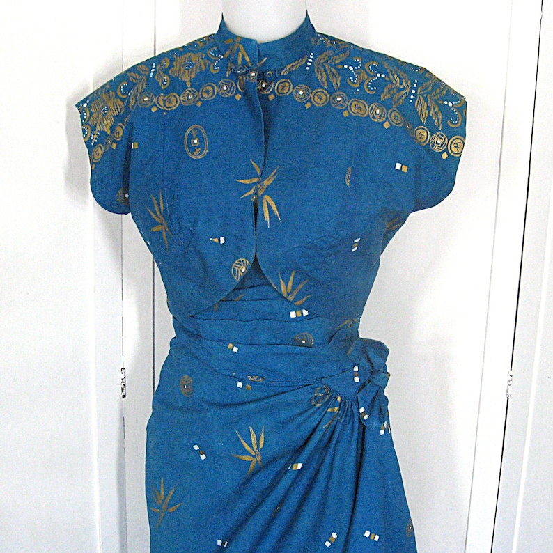 May include: A teal dress and bolero jacket set. The garments are adorned with gold leaf and geometric patterns. The dress has a wrap skirt and high neckline. The bolero features a mandarin collar and short sleeves. The dress is a vintage style.
