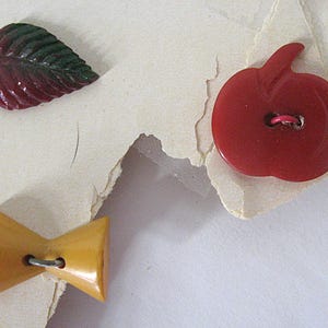 May include: Three decorative buttons including a dark red and green leaf-shaped button, a solid red apple-shaped button with a central hole, and a mustard yellow bow-tie shaped button with a central hole, all featuring smooth surfaces.