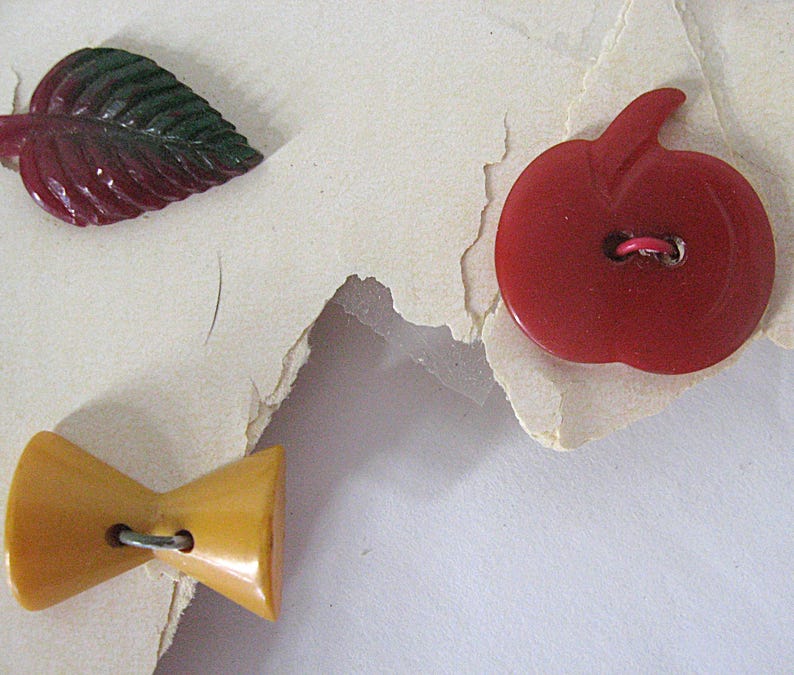 May include: Three decorative buttons including a dark red and green leaf-shaped button, a solid red apple-shaped button with a central hole, and a mustard yellow bow-tie shaped button with a central hole, all featuring smooth surfaces.