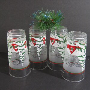 May include: Set of four frosted glass tumblers with a winter scene design. Each glass features a white house with a red roof, green pine trees, and a snowy background. The glasses have a red and green band around the top.
