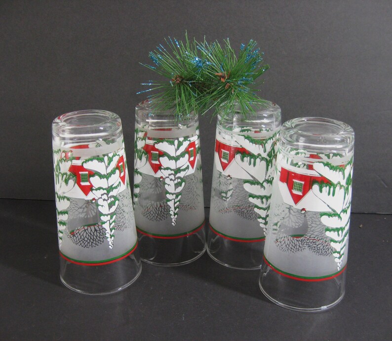 May include: Set of four frosted glass tumblers with a winter scene design. Each glass features a white house with a red roof, green pine trees, and a snowy background. The glasses have a red and green band around the top.