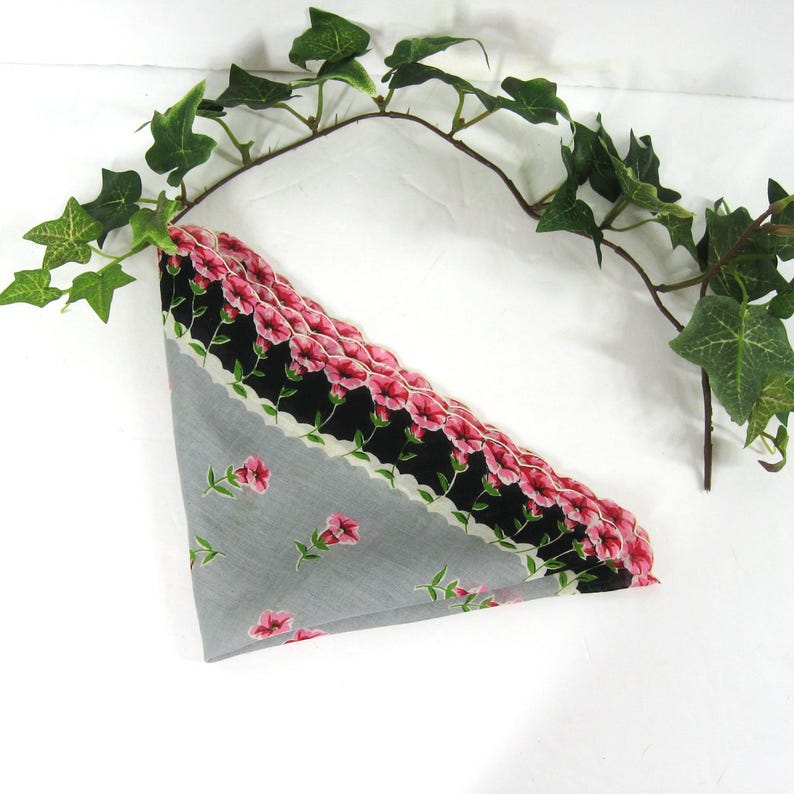 Pode incluir: Um len&ccedil;o cinza com uma borda festonada preta e branca e flores cor-de-rosa. O len&ccedil;o est&aacute; dobrado em forma de tri&acirc;ngulo.
