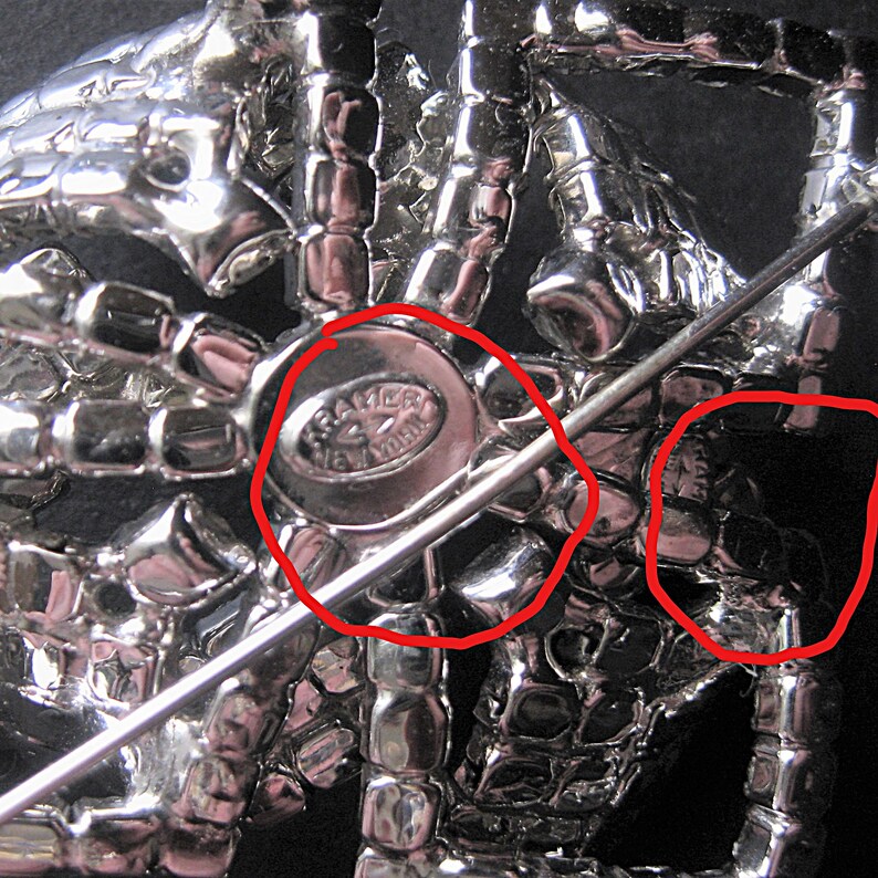 May include: A silver-toned brooch in the shape of a spider with rhinestones. The back of the brooch has a metal pin and a maker's mark that reads "Brazer New York".