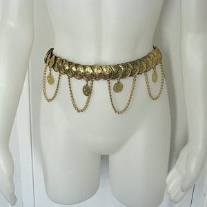 May include: A gold-colored coin belt with dangling chains and coins. The belt is displayed on a mannequin. The belt is made of gold-colored metal and features a series of round coins with decorative patterns. Chains hang from the belt, each with a small coin at the end.