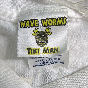 May include: White t-shirt tag with yellow and black text that reads "WAVE WORMS TIKI MAN". The tag also has a black and yellow tiki mask graphic. The tag has additional text that reads "100% COTTON KNIT IN U.S.A. ASSEMBLED IN HONDURAS".