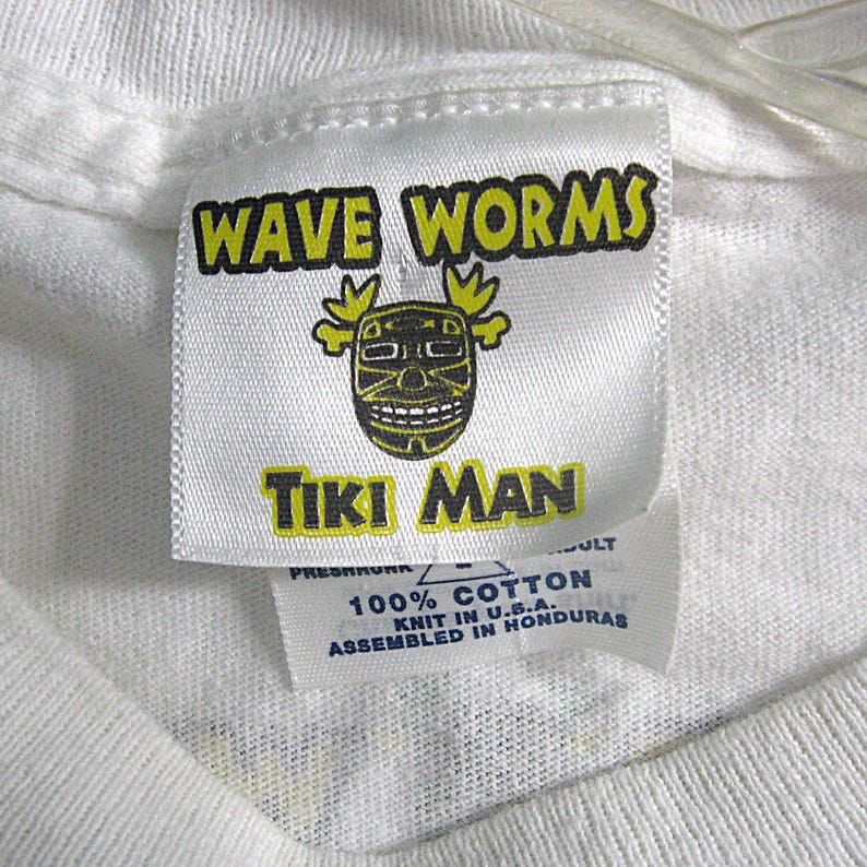 May include: White t-shirt tag with yellow and black text that reads "WAVE WORMS TIKI MAN". The tag also has a black and yellow tiki mask graphic. The tag has additional text that reads "100% COTTON KNIT IN U.S.A. ASSEMBLED IN HONDURAS".