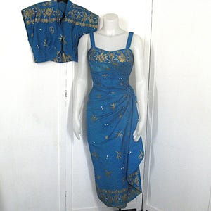 May include: A teal dress and matching bolero jacket set. The dress has a draped design and spaghetti straps. Both items are decorated with gold embroidery and small white accents. The set is displayed on a mannequin.