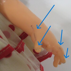 May include: A peach-toned plastic doll arm and hand featuring several dark ink marks on the skin, highlighted by four bright blue arrows pointing to the specific areas of discoloration on the wrist, thumb, and fingers, worn with a white dress.