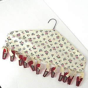 May include: A vintage clothes hanger with a floral fabric cover and red clips. The fabric is cream-colored with a pattern of pink and blue flowers. The clips are attached with cream-colored ribbons.