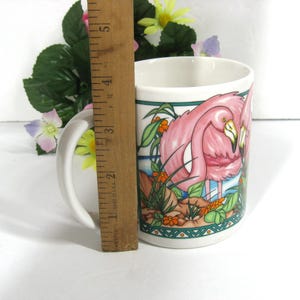 May include: White ceramic mug with a pink flamingo design. The mug is 10 cm tall and features a green and blue border around the image.