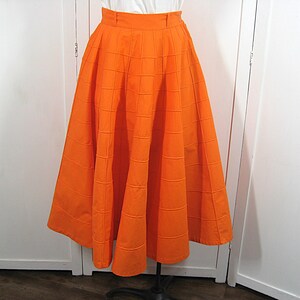 May include: A bright orange full skirt with a box pleat design. The skirt is made of a textured fabric and has a fitted waistband.