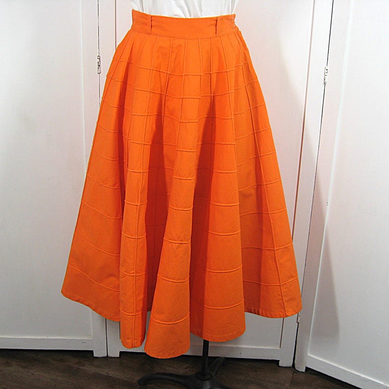 May include: A bright orange full skirt with a box pleat design. The skirt is made of a textured fabric and has a fitted waistband.