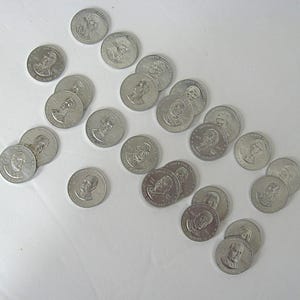 May include: A collection of silver-coloured commemorative coins featuring portraits of historical figures. The coins are scattered across a white surface, with various designs and inscriptions visible. The coins appear to be in good condition.