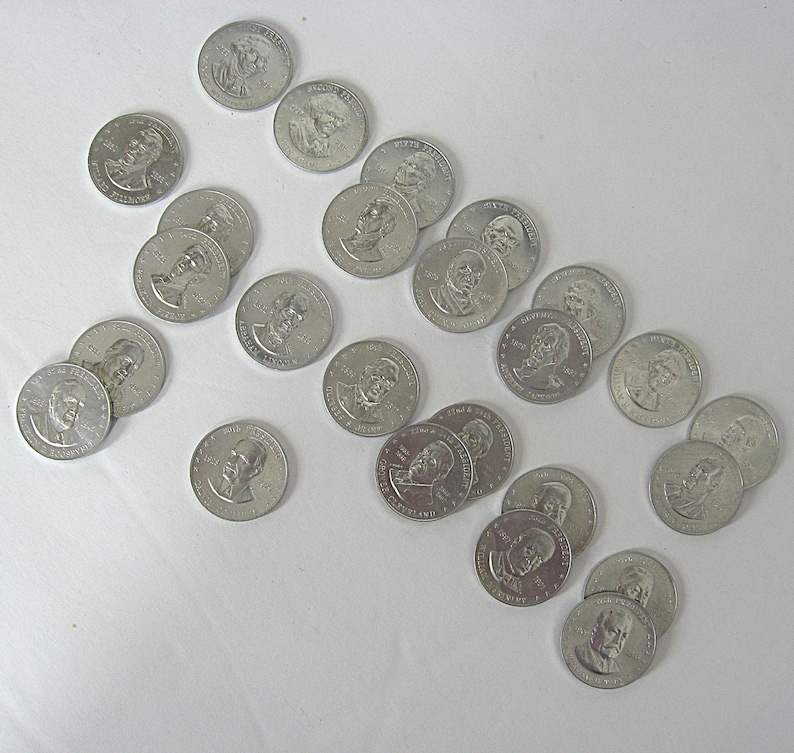 May include: A collection of silver-coloured commemorative coins featuring portraits of historical figures. The coins are scattered across a white surface, with various designs and inscriptions visible. The coins appear to be in good condition.