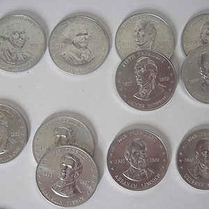 May include: A collection of silver-coloured commemorative coins featuring portraits of U.S. presidents. The coins display the names and terms of office of presidents such as George Washington, John Adams, and Abraham Lincoln.