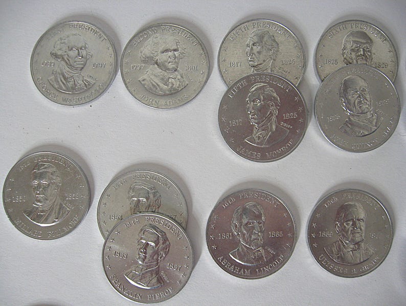 May include: A collection of silver-coloured commemorative coins featuring portraits of U.S. presidents. The coins display the names and terms of office of presidents such as George Washington, John Adams, and Abraham Lincoln.