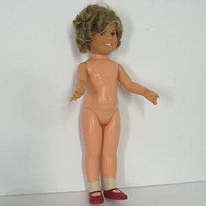 May include: A doll with light skin, short curly brown hair, and a smiling expression. The doll has a peach-colored body and is wearing white socks and dark red shoes. The doll is standing in an upright position with arms extended outward.