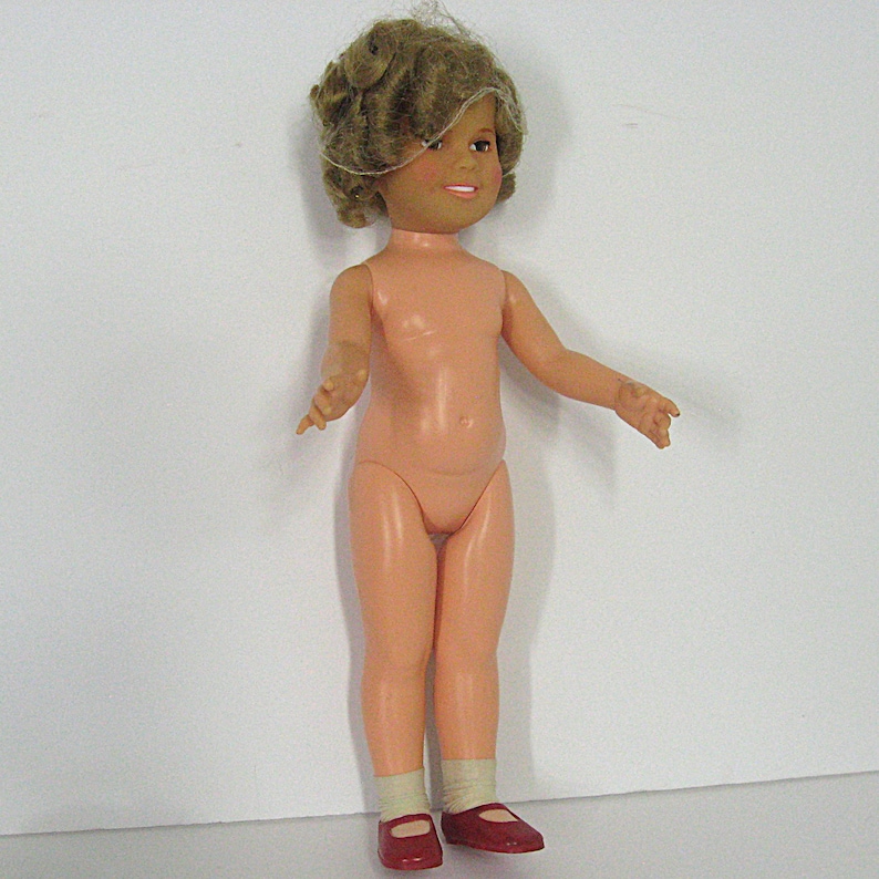 May include: A doll with light skin, short curly brown hair, and a smiling expression. The doll has a peach-colored body and is wearing white socks and dark red shoes. The doll is standing in an upright position with arms extended outward.