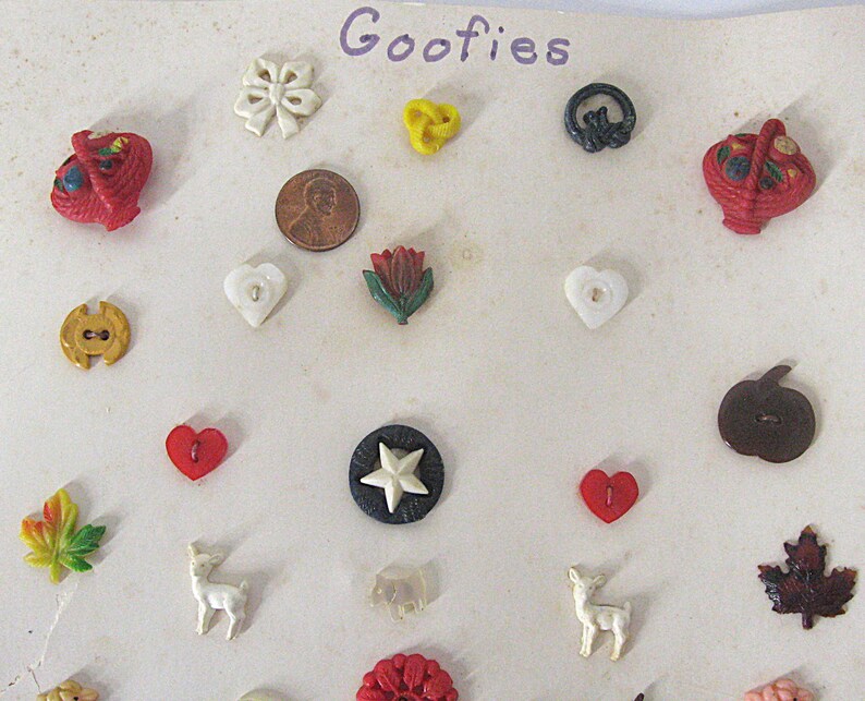 May include: A collection of assorted decorative buttons labeled Goofies, including shapes like hearts, leaves, stars, deer, and baskets in various colors. A copper penny is included for scale, measuring approximately 0.75 inches in diameter.