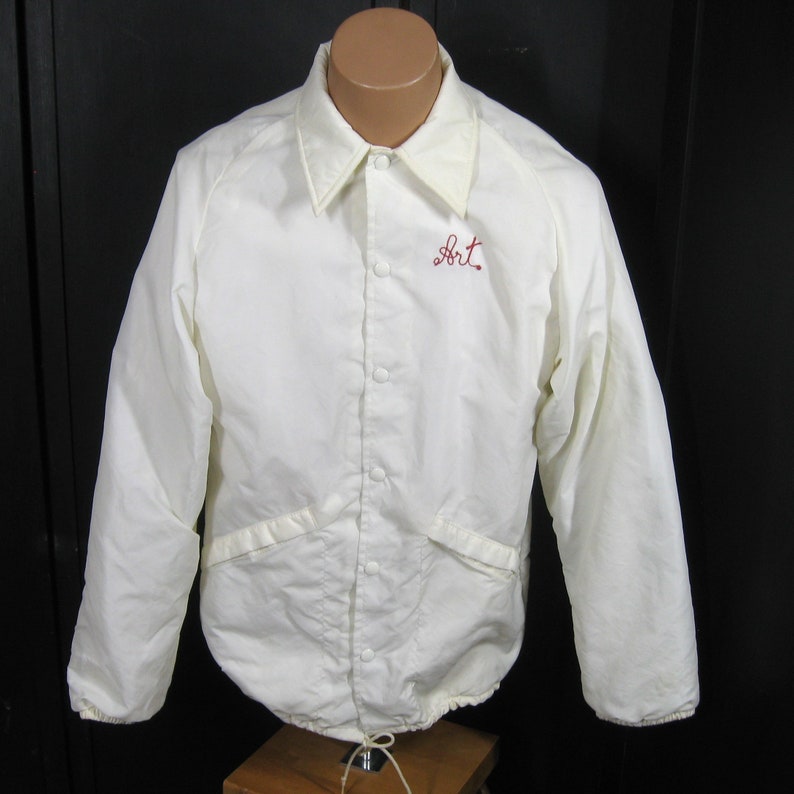 Pode incluir: Casaco vintage branco com um "Art" bordado em vermelho no peito esquerdo. O casaco tem fecho de bot&otilde;es, mangas compridas e dois bolsos.