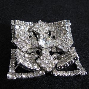May include: A silver-tone brooch featuring a large clear rhinestone at its centre, surrounded by smaller rhinestones. The brooch is shaped like a flower with multiple layers of rhinestones.