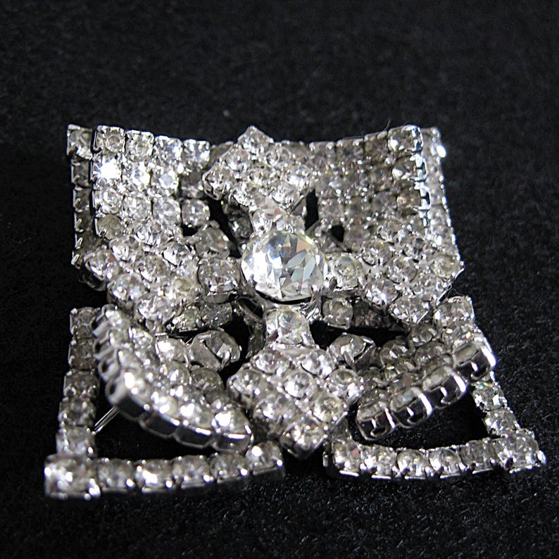 May include: A silver-tone brooch featuring a large clear rhinestone at its centre, surrounded by smaller rhinestones. The brooch is shaped like a flower with multiple layers of rhinestones.