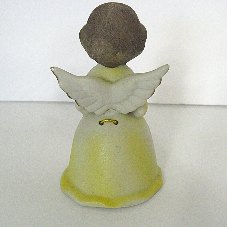 May include: A ceramic angel figurine viewed from the back, featuring a yellow dress and white wings with gold trim. The angel has brown hair and a small gold accent at the base of the wings. The figurine is on a white surface.