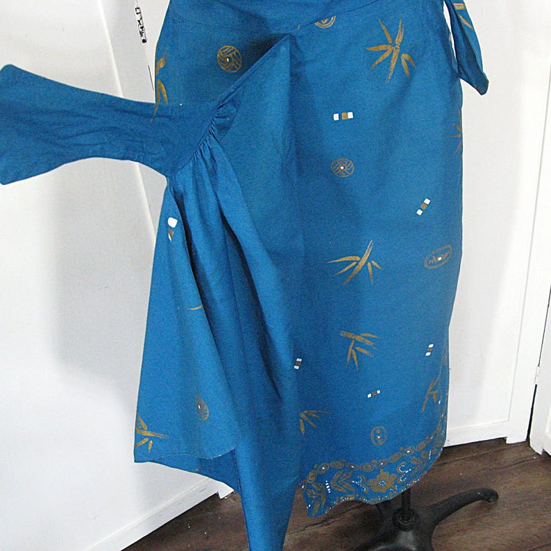 May include: A teal wrap skirt with gold bamboo and circular patterns, plus small white and gold accents. The bottom hem has a decorative border. The skirt is displayed on a black metal stand. The fabric appears to be a lightweight material.
