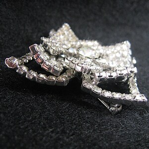 May include: A silver-toned brooch with a bow design, featuring a cluster of clear rhinestones. The brooch has a pin and catch closure.