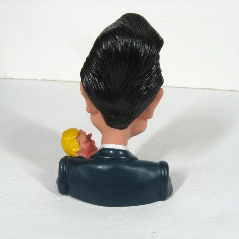 May include: A caricature bust of a person with black hair and a blue suit holding a smaller caricature bust of a person with yellow hair.