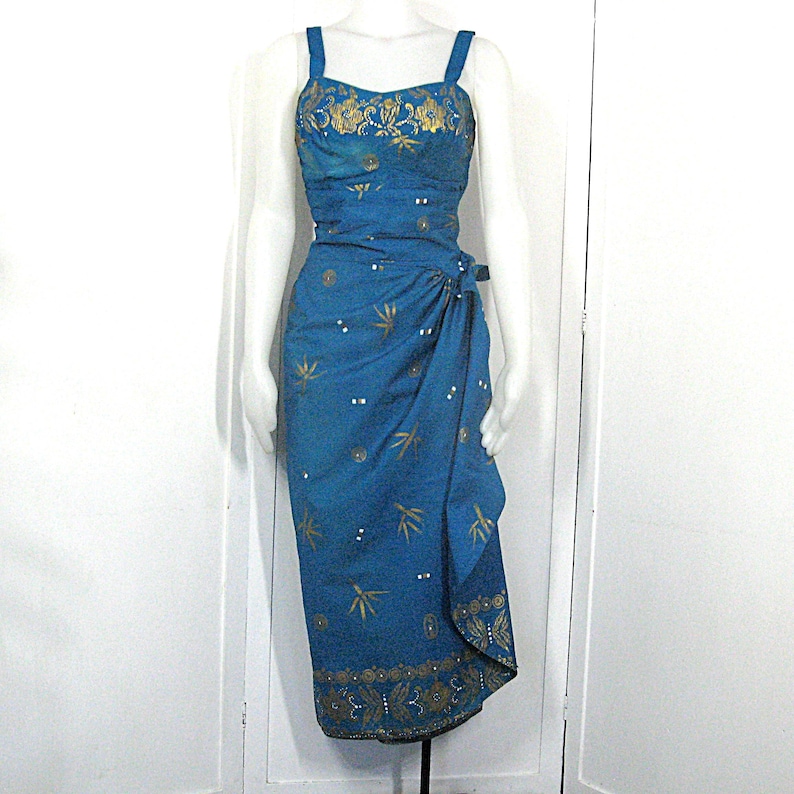May include: A teal dress with a wrap skirt and spaghetti straps. The bodice and skirt feature gold floral embroidery and small white details. The dress is displayed on a mannequin.