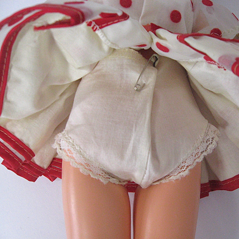 May include: A doll wearing white underpants with lace trim and a safety pin attached to the front, underneath a white skirt featuring a red polka dot pattern and a red hem. The doll has smooth, light-toned legs visible below the undergarments.