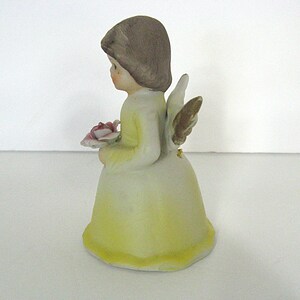 May include: A ceramic figurine of an angel, wearing a yellow dress and holding a pink rose. The angel has brown hair and gold-colored wings. The figurine is a decorative item.
