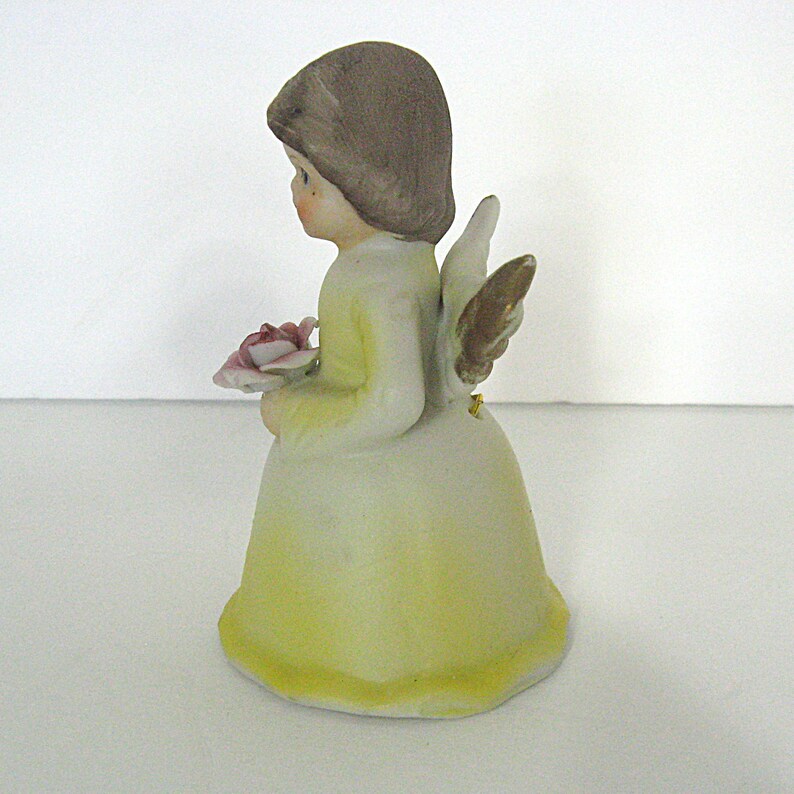 May include: A ceramic figurine of an angel, wearing a yellow dress and holding a pink rose. The angel has brown hair and gold-colored wings. The figurine is a decorative item.