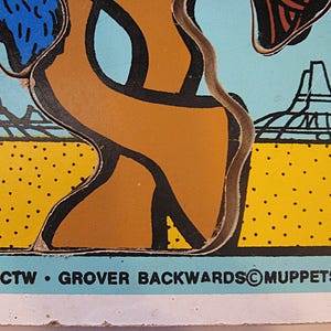 Pode incluir: Uma pe&ccedil;a de quebra-cabe&ccedil;a vintage com fundo azul claro, uma paisagem des&eacute;rtica amarela, formas abstratas marrons e contornos pretos. O texto diz: 1976 CTW GROVER BACKWARDS MUPPETS, INC. e MADE IN U.S.A. impresso na borda inferior.