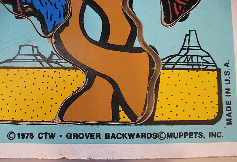 Pode incluir: Uma pe&ccedil;a de quebra-cabe&ccedil;a vintage com fundo azul claro, uma paisagem des&eacute;rtica amarela, formas abstratas marrons e contornos pretos. O texto diz: 1976 CTW GROVER BACKWARDS MUPPETS, INC. e MADE IN U.S.A. impresso na borda inferior.