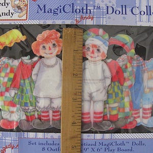 May include: A MagiCloth doll collection set featuring paper dolls with outfits. The set includes two dolls with red hair and painted faces, along with several outfits in various colors and patterns. The packaging includes the text "MagiCloth" and "Andy".