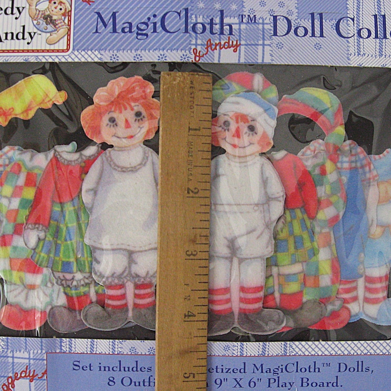 May include: A MagiCloth doll collection set featuring paper dolls with outfits. The set includes two dolls with red hair and painted faces, along with several outfits in various colors and patterns. The packaging includes the text "MagiCloth" and "Andy".