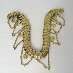 May include: A gold-colored belly dance belt with rows of decorative coins. The belt features dangling chains and smaller coins, creating a flowing, ornate design. The coins have a detailed floral pattern.