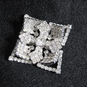 May include: A silver-toned brooch with a square shape and a large, clear rhinestone in the centre. The brooch is made of many smaller rhinestones arranged in a geometric pattern.