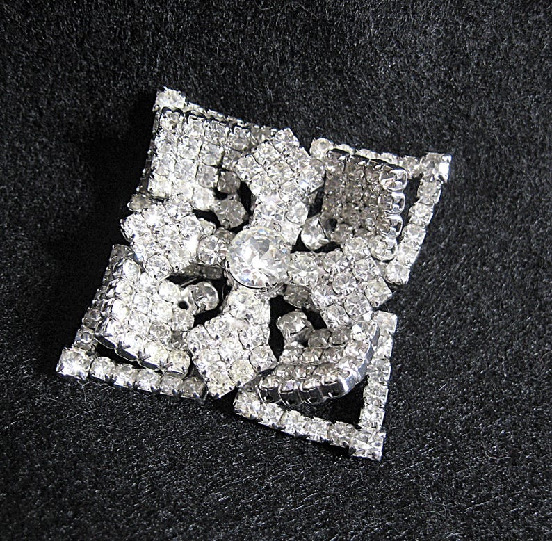 May include: A silver-toned brooch with a square shape and a large, clear rhinestone in the centre. The brooch is made of many smaller rhinestones arranged in a geometric pattern.